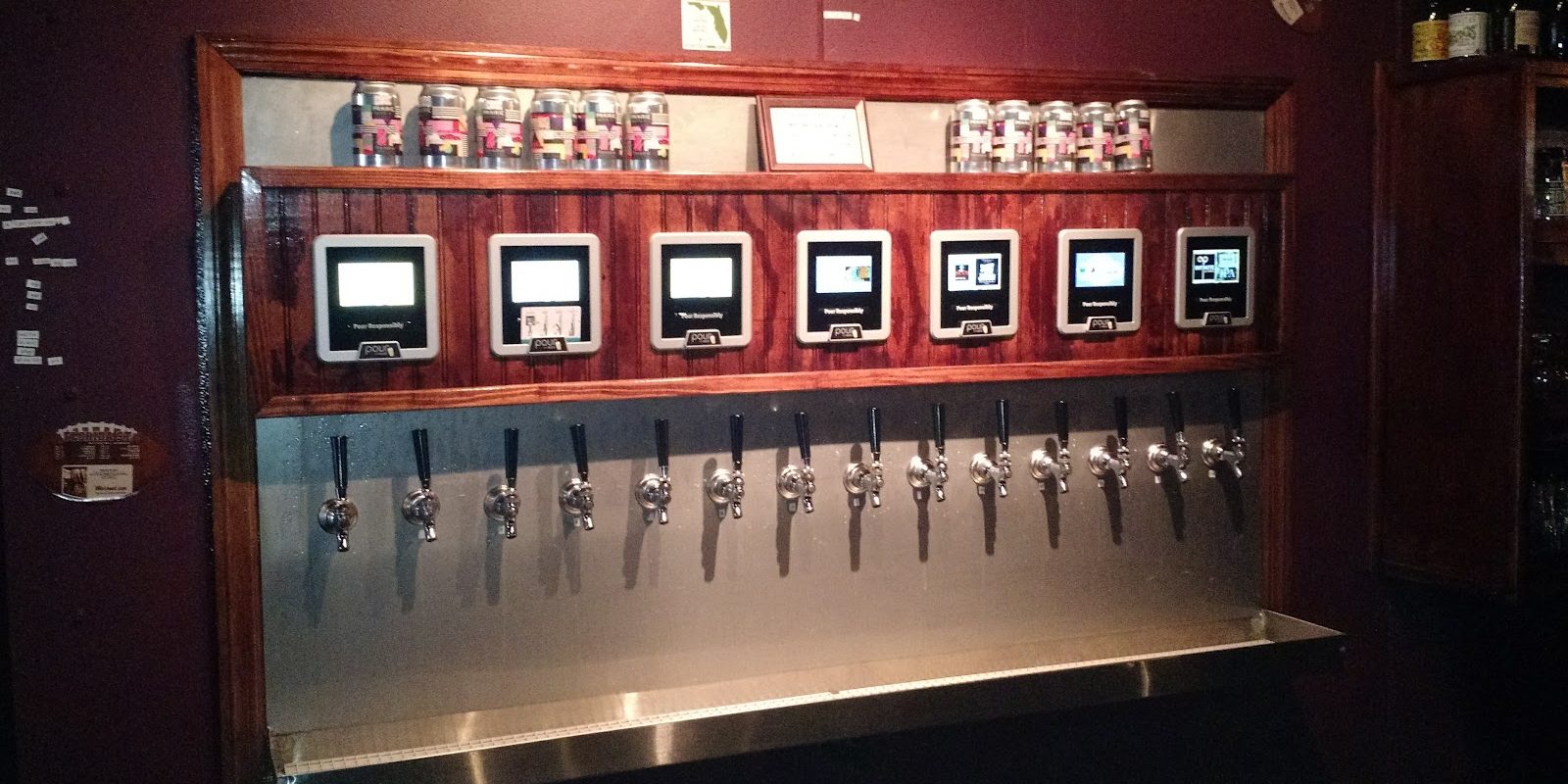 A long stainless tap wall framed in red wood, a row of black tap handles under seven small touchscreens, labelled cans on the shelf above.